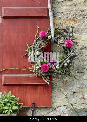 Corona floreale di Pasqua fatta di ranunculus e rami di alberi da frutta con uova di quaglia, appeso su un serramento di legno di fronte a muratura di pietra di cava Foto Stock