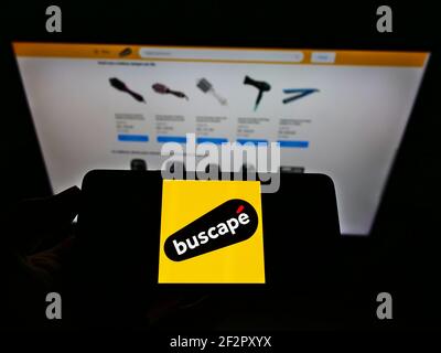 Persona che tiene il telefono cellulare con il logo aziendale del negozio online brasiliano Buscapé sullo schermo di fronte alla pagina web aziendale. Focus sul display del cellulare. Foto Stock