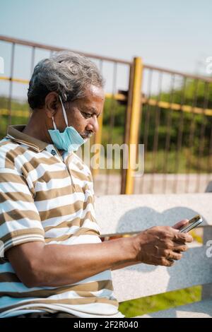Uomo anziano con maschera medica sotto la mascella utilizzando smartphone al parco - concetto di uso improprio maschera a causa di pandemie coronavirus covid-19. Foto Stock