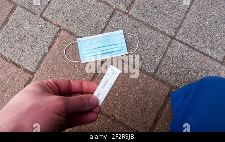 Cassetta bianca per analisi corona con risultato negativo covid-19. La maschera medica per bocca e naso giace su ciottoli sporchi. Ginocchio con pantaloni da lavoro. Da a. Foto Stock