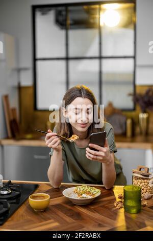 Bella donna in casual t-shirt verde tiene un cucchiaio con granola in mano e legge al telefono durante una mattina in cucina a casa Foto Stock
