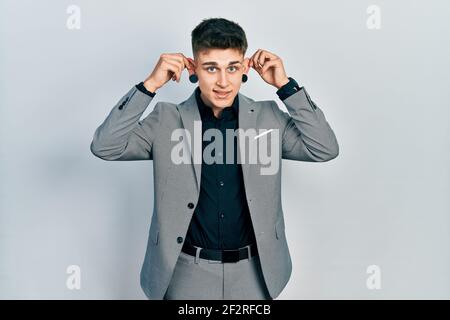 Ragazzo caucasico giovane con le orecchie dilatazione indossare giacca da lavoro sorridente tirando le orecchie con le dita, gesto divertente. Problema di audizione Foto Stock