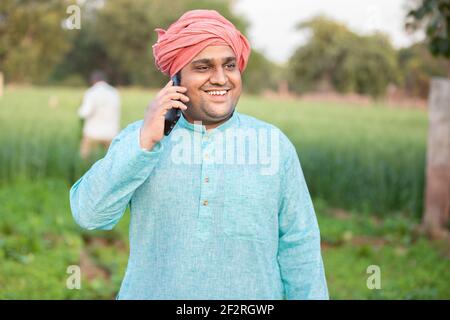 Giovane lavoratore contadino indiano felice che parla al telefono mentre si trova in campo verde, agricoltura e concetto di tecnologia, maschio indossando kurta tradizionale ou Foto Stock