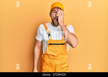 Giovane bell'uomo che indossa uniforme di handyman su sfondo giallo sbadigli stanco coprendo metà faccia, occhio e bocca con mano. Faccia fa male. Foto Stock