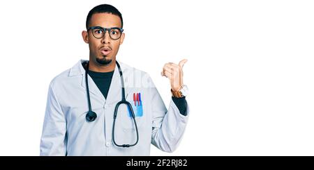 Giovane afroamericano uomo indossando uniforme medico e stetoscopio sorpreso puntando con la mano dito a lato, bocca aperta stupito espressione. Foto Stock