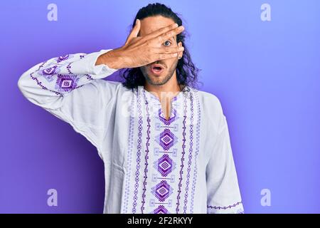 Giovane bell'uomo con capelli lunghi che indossa camicia bohémien e hippie che spazza in shock coprendo viso e occhi con la mano, guardando attraverso le dita con em Foto Stock