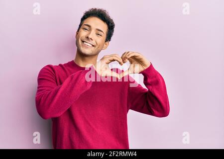 Bell'uomo ispanico che indossa abiti casual su sfondo rosa sorridente in amore facendo il simbolo del cuore forma con le mani. Concetto romantico. Foto Stock