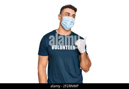 Giovane ispanico che indossa maschera medica e t shirt volontaria sorridente con volto felice guardando e puntando al lato con il pollice in su. Foto Stock