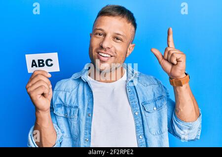 Bell'uomo del muscolo che tiene le lettere abc sulla carta sorridente con un'idea o una domanda che punta il dito con il volto felice, il numero uno Foto Stock