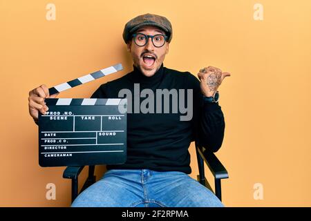 Bell'uomo con tatuaggi che tengono la clapboard del video film seduto sopra sedia director che punta il pollice verso il lato sorridendo felice a bocca aperta Foto Stock