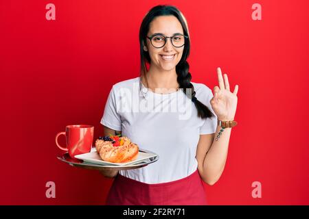 Giovane donna ispanica che indossa grembiule cameriera reggendo vassoio con colazione facendo ok segno con le dita, sorridente amichevole gesturing eccellente simbolo Foto Stock