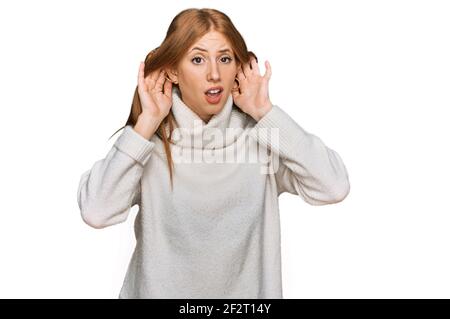 Giovane donna irlandese che indossa un maglione invernale casual cercando di sentire entrambe le mani sul gesto dell'orecchio, curiosa per pettegolezzi. Problemi di udito, sordi Foto Stock