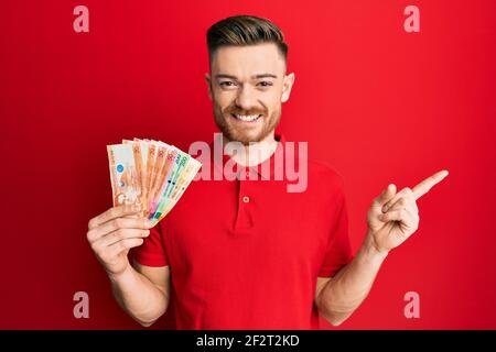 Giovane uomo rosso che tiene le banconote del peso filippino sorridente che indica felice con la mano e il dito di lato Foto Stock