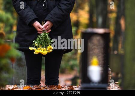 Donna lutto che tiene i fiori in mano e in piedi alla tomba nel cimitero. Tristezza durante i funerali Foto Stock