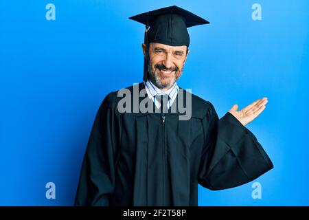 Uomo ispanico di mezza età che indossa un cappuccio di graduazione e la cerimonia robe sorridente allegro presentazione e puntando con palmo di mano guardando la fotocamera. Foto Stock
