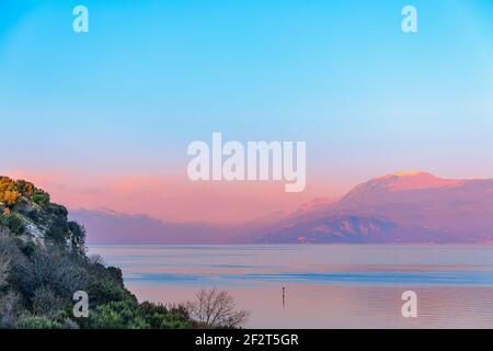 Splendida vista sul tranquillo Lago di Garda e sul Monte Balbo, con i raggi del tramonto e la nebbia serale. Periodo invernale. Vista dalla penisola di Sirmione, Ital Foto Stock
