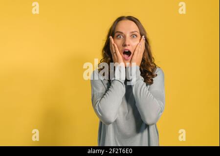 Scioccato e stupito giovane attraente donna guarda la telecamera isolata sul giallo, una ragazza caucasica maledetta urla sorpreso, tenendo guance, vendite incredibili o affare Foto Stock