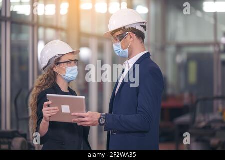 Ritratto di due macchinisti è in piedi con tablet e casco di sicurezza di fronte alla parete di vetro della fabbrica industriale. Foto Stock