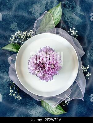 Composizione della stagione primaverile con un piatto bianco e fiori su sfondo blu. Romantico tavolo floreale con grandi foglie e gypsophila. Weddin Foto Stock