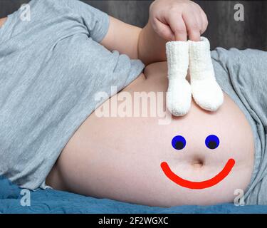 Solo per divertimento. Una donna incinta che tiene il bambino ha lavorato i calzini piccoli nelle sue mani ed un sorriso felice tirato sul suo ventre. Concetto allegro felice di gravidanza. Foto Stock