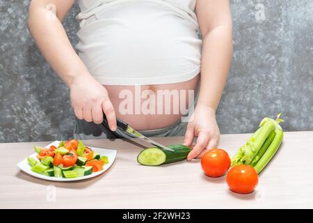 Donna incinta taglio cetriolo per insalata fresca verde, donna prepara gustosa cena biologica a casa, nutrizione sana per futura madre. Concetto di h Foto Stock