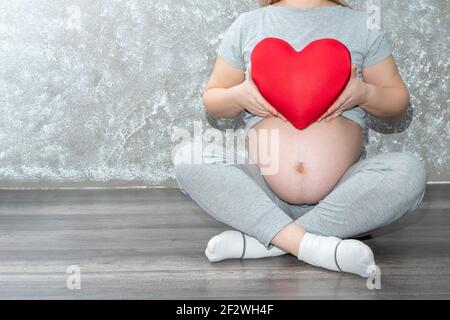 Concetto di gravidanza, maternità, persone, amore e aspettativa - primo piano di una donna incinta che tiene un cuscino a forma di cuore rosso a casa. Una donna incinta Foto Stock