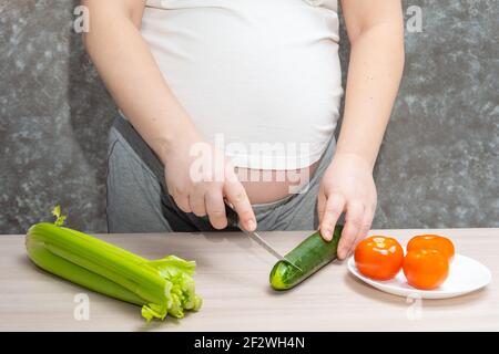 Donna incinta taglio cetriolo per insalata fresca verde, donna prepara gustosa cena biologica a casa, nutrizione sana per futura madre. Concetto di h Foto Stock