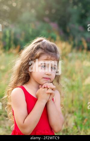 Ritratto di carina bambina che prega nel parco, bambino che tiene le mani insieme, closeup espressione. Religione fede e credere concetto Foto Stock