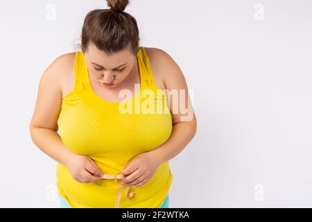 Foto ritagliata di una donna più grande in una T-shirt luminosa che misura il suo ventre con un centimetro. Sovrappeso e obesità su sfondo bianco. Foto Stock