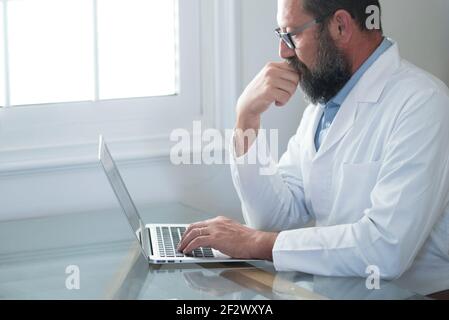 Adulto maturo caucasico maschio medico in uniforme medica bianca sedersi alla scrivania in ufficio consultare il cliente su portatile online, uomo GP o medico riempire paziente me Foto Stock