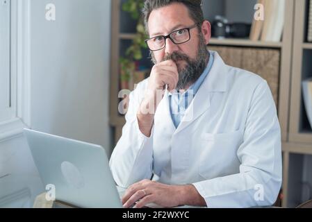 Adulto maturo caucasico maschio medico in uniforme medica bianca sedersi alla scrivania in ufficio consultare il cliente su portatile online, uomo GP o medico riempire paziente me Foto Stock