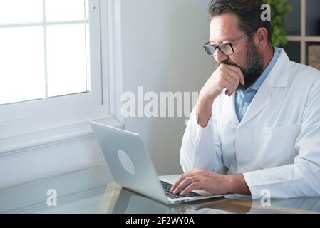 Medico maturo seduto in ufficio. Per adulti, scrivete con un cappotto bianco e lavorate alla scrivania con un computer portatile. Consulenza medica professionale e trattamento in moderno Foto Stock