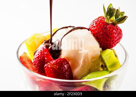 Gelato alla vaniglia in un bicchiere con fragola kiwi e. morde di arance e cioccolato fondente che vengono versati dalla parte superiore isolato su sfondo bianco in stu Foto Stock
