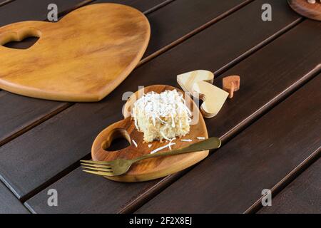 Torta di cocco con guarnizione di cocco grattugiata accanto a una forchetta di rame. Su una piastra a forma di cuore con diversi cuori di legno accanto ad essa. Foto Stock