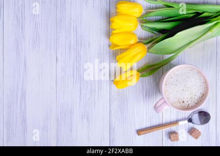 Vista dall'alto di una composizione di un bouquet di tulipani gialli, una tazza rosa con cappuccino con un cucchiaio e zucchero su uno sfondo di legno chiaro. Messa a fuoco selettiva Foto Stock