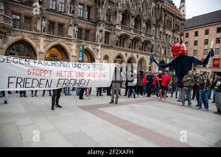 Demonstrant*innen halten trasparente: ' Gegn Faschismus und Diktatur stopt Hetze und Spaltung Frieden Freiheit ' und rechts daneben ist eine Teufelsfigur, die eine Impfspritze hält. Am 13. März 2021 versammelten sich tausende Menschen in München, um gegen sämtliche Covid Maßnahmen zu demonstrieren. Eine Demonstration vom Königsplatz startend wurde vom Versammlungsleiter Markus Haintz abgesagt, weil die Polizei die Teilnehmerbeschränkung durchsetzte. Die Kundgebung am MaxMonument überschritt die 500 erlaubten Teilnehmer*innen deutlich. Viele trugen keine Maske oder trugen sie nicht richtig. Foto Stock