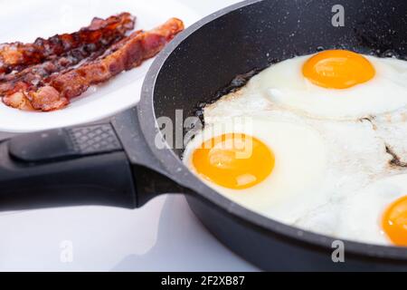 Uova fritte e pancetta cucinando in una padella o in una padella. Idea per la colazione del mattino. Tuorli gialli di uova e fette rosse di pancetta fritta Foto Stock