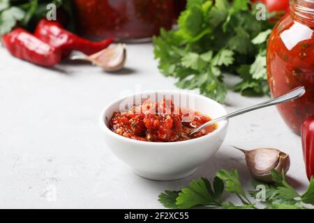 Adjika armena di pepe rosso, pomodori, aglio, coriandolo e prezzemolo in recipiente e vasetti, nonché ingredienti freschi su tavola leggera. Foto Stock