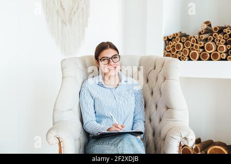 Ritratto di sorridente psicologa caucasica seduta in poltrona sul posto di lavoro, tenendo una cartella con carte per appunti... Foto Stock