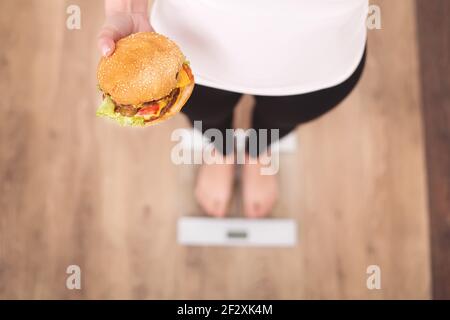 Concetto di dieta e fast food. Donna sovrappeso in piedi su bilancia di pesatura Holding Burger ( Hamburger ). Cibo spazzatura malsano. Dieta, stile di vita. Peso Foto Stock