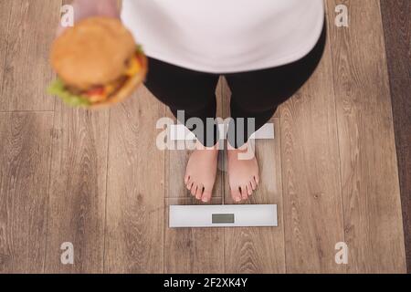 Concetto di dieta e fast food. Donna sovrappeso in piedi su bilancia di pesatura Holding Burger Hamburger . Cibo spazzatura malsano. Dieta, stile di vita. Peso Foto Stock