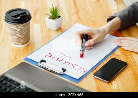 Ricerca di lavoro. Femmina scrive a mano riprendere con il rossetto sul foglio di carta bianco. Ufficio in legno scrivania con laptop, smartphone e i materiali di consumo Foto Stock