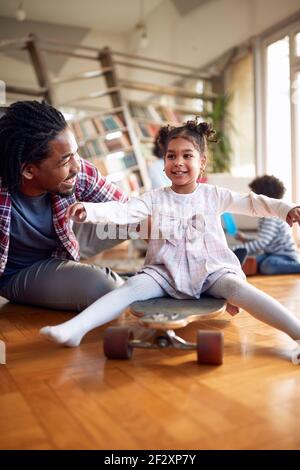 Una bambina ama guidarla sullo skateboard in un'atmosfera divertente a casa. Famiglia, insieme, amore, tempo di gioco Foto Stock