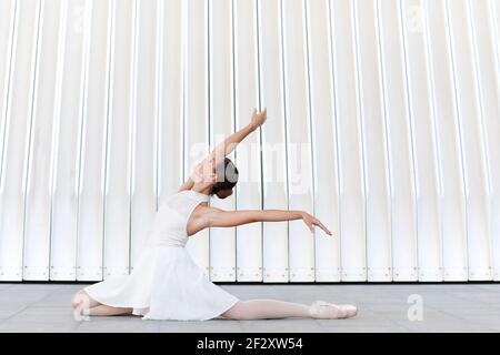 Vista laterale della giovane ballerina aggraziata in abito bianco seduto sul passerella mentre si esegue il balletto posa e guardando in su Foto Stock