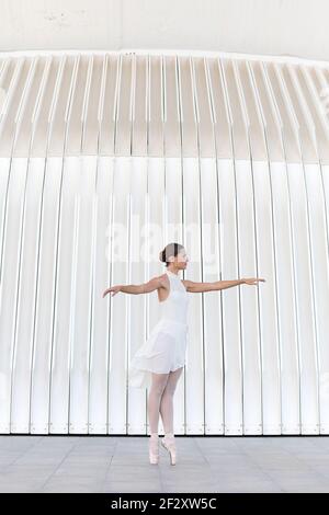 Giovane ballerina femminile in punta con scarpe pointe gambe rialzate e braccia danzanti su pavimenti piastrellati all'aperto Foto Stock
