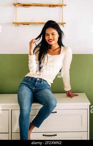 Giovane donna ispanica allegra che indossa jeans seduti sul bancone in una stanza moderna e luminosa e che guarda la macchina fotografica Foto Stock