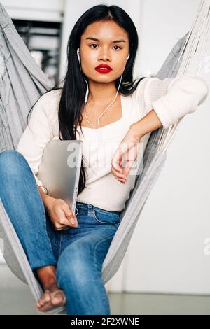 Giovane donna premurosa con lunghi capelli scuri che tiene un portatile con auricolari seduti in amaca al chiuso guardando lontano Foto Stock