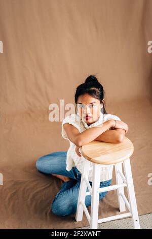 Corpo pieno di giovane donna ispanica sicura che indossa abiti casual seduta sul pavimento appoggiata su sgabello di legno in studio Foto Stock