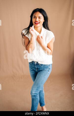 Femmina etnico soddisfatto in abito casual con sorriso toothy guardando in studio, mentre si sta in piedi su sfondo marrone Foto Stock