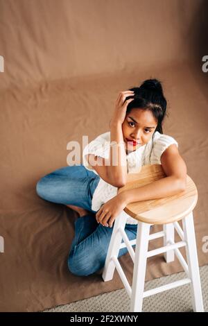 Corpo pieno di giovane donna ispanica sicura che indossa abiti casual seduta sul pavimento appoggiata su sgabello di legno in studio Foto Stock